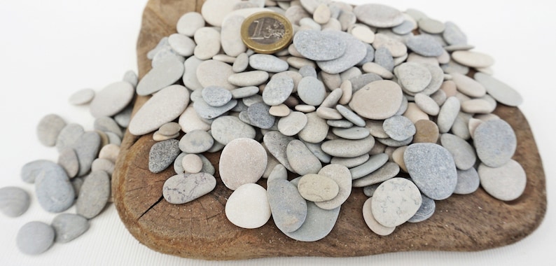 May include: A pile of smooth, gray and white pebbles of various sizes on a wooden surface. A Euro coin is included for scale.