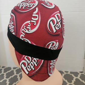 May include: A skull cap featuring a repeating Dr Pepper soda logo pattern in white and red on a dark red background. The cap includes a solid black horizontal band across the lower section and is constructed with visible stitched seams throughout.
