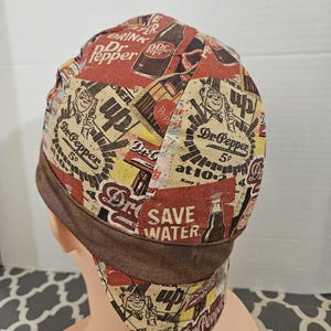 May include: A fabric cap featuring a vintage-style Dr. Pepper soda advertisement print in shades of red, brown, and cream. The design includes text such as Drink Dr. Pepper, 5 cents, at 10-2, and Save Water, with a solid brown band around the base.