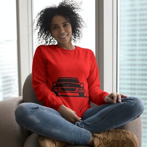 May include: A woman wearing a red sweatshirt with a black silhouette of a car on the front. She is sitting on a gray armchair with her legs crossed.