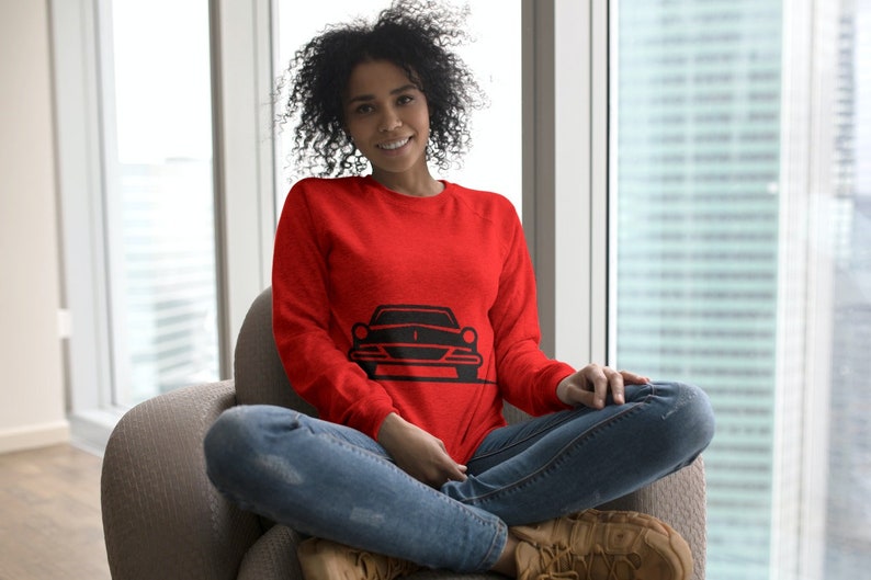 May include: A woman wearing a red sweatshirt with a black silhouette of a car on the front. She is sitting on a gray armchair with her legs crossed.