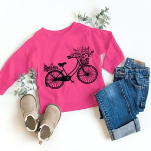 May include: A pink long-sleeved shirt with a black graphic of a bicycle with a basket of flowers. The shirt is paired with blue jeans and brown boots.