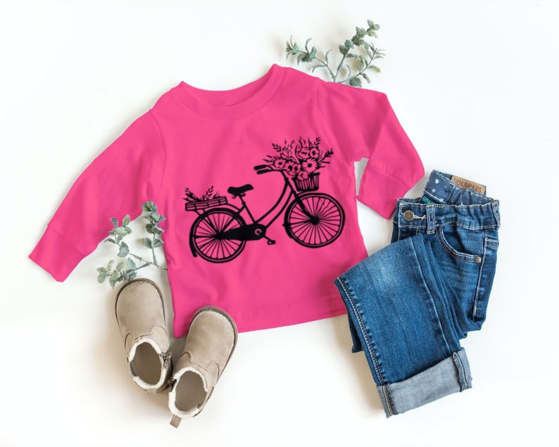 May include: A pink long-sleeved shirt with a black graphic of a bicycle with a basket of flowers. The shirt is paired with blue jeans and brown boots.