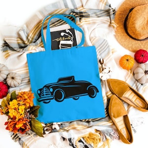 May include: A blue tote bag with a black silhouette of a vintage car printed on the front. The bag is surrounded by fall-themed decor, including pumpkins, leaves, and a straw hat.