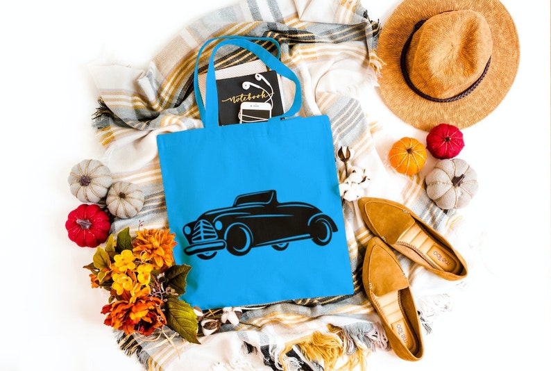 May include: A blue tote bag with a black silhouette of a vintage car printed on the front. The bag is surrounded by fall-themed decor, including pumpkins, leaves, and a straw hat.