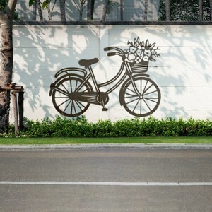 May include: A black silhouette of a bicycle with a basket of flowers painted on a white wall. The bicycle is in front of a green bush and a tree.
