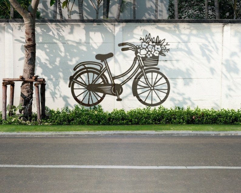 May include: A black silhouette of a bicycle with a basket of flowers painted on a white wall. The bicycle is in front of a green bush and a tree.
