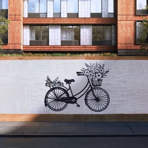 May include: A black bicycle with a basket of flowers on the handlebars. The bicycle is on a white background with a brick building in the background.