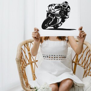 May include: A woman holds a white poster in front of her face. The poster features a black and white silhouette of a motorcycle racer riding a motorcycle.
