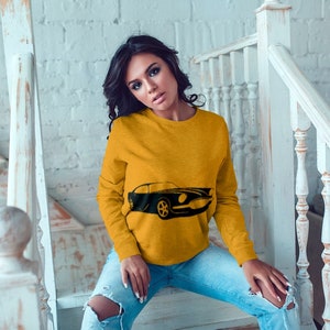 May include: A woman wearing a yellow sweatshirt with a black silhouette of a classic car printed on the front. She is sitting on a set of stairs with a white railing.