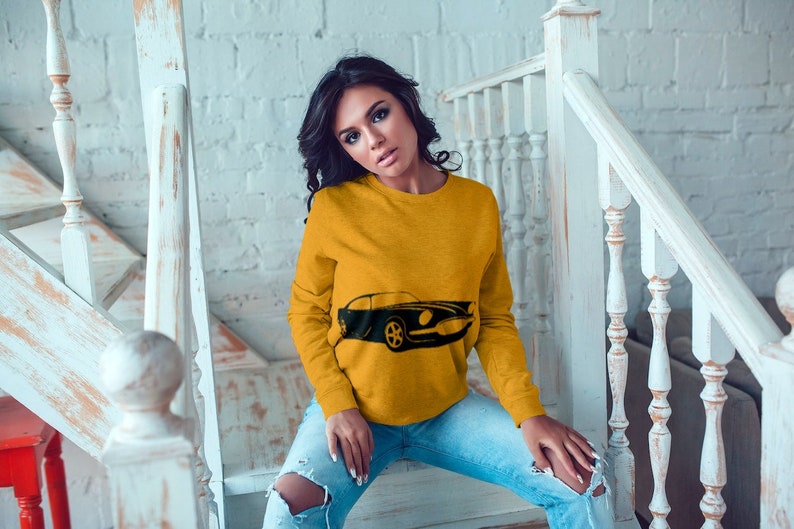 May include: A woman wearing a yellow sweatshirt with a black silhouette of a classic car printed on the front. She is sitting on a set of stairs with a white railing.