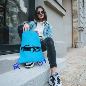 May include: A woman wearing sunglasses sits on a step with a blue drawstring bag featuring a black car graphic. The bag has blue drawstrings.