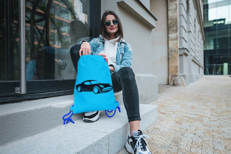 May include: A woman wearing sunglasses sits on a step with a blue drawstring bag featuring a black car graphic. The bag has blue drawstrings.