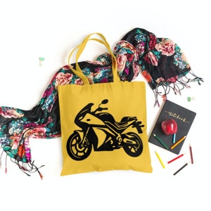 May include: A yellow tote bag with a black silhouette of a motorcycle on the front. The bag is sitting on a white surface with a floral scarf and a notebook.