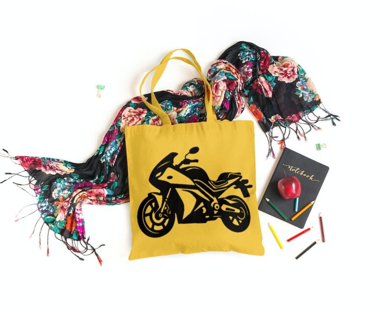 May include: A yellow tote bag with a black silhouette of a motorcycle on the front. The bag is sitting on a white surface with a floral scarf and a notebook.