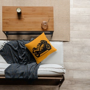 May include: A yellow throw pillow with a black silhouette of a motorcycle on it. The pillow is on a white couch with a gray throw blanket.