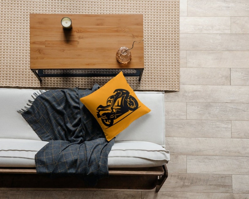 May include: A yellow throw pillow with a black silhouette of a motorcycle on it. The pillow is on a white couch with a gray throw blanket.