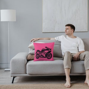 May include: A pink throw pillow with a black silhouette of a motorcycle on it. The pillow is on a gray couch with a person sitting on it.