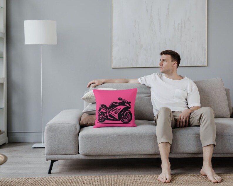May include: A pink throw pillow with a black silhouette of a motorcycle on it. The pillow is on a gray couch with a person sitting on it.