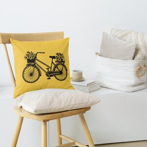 May include: A yellow throw pillow with a black silhouette of a bicycle with flowers in the basket. The pillow is on a wooden chair with a white background.