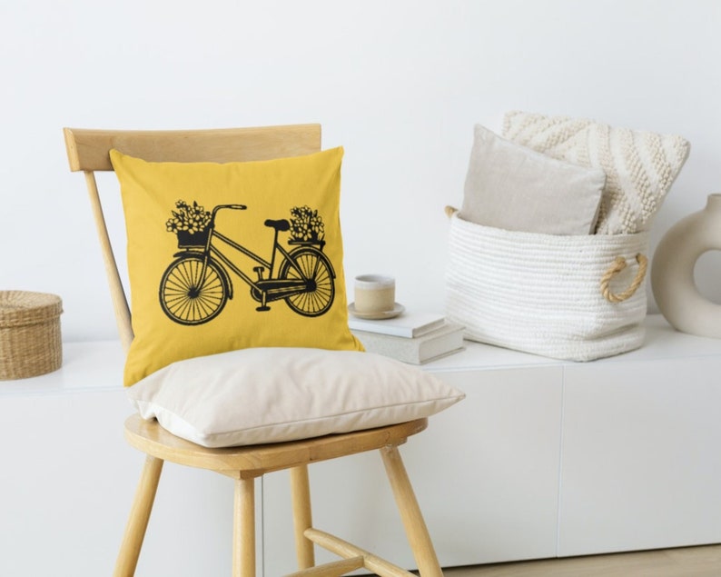 May include: A yellow throw pillow with a black silhouette of a bicycle with flowers in the basket. The pillow is on a wooden chair with a white background.
