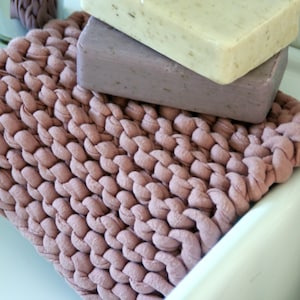 May include: A pink knitted soap dish with three bars of soap on top. The soap dish has a long, thin strap that can be used to hang it up.