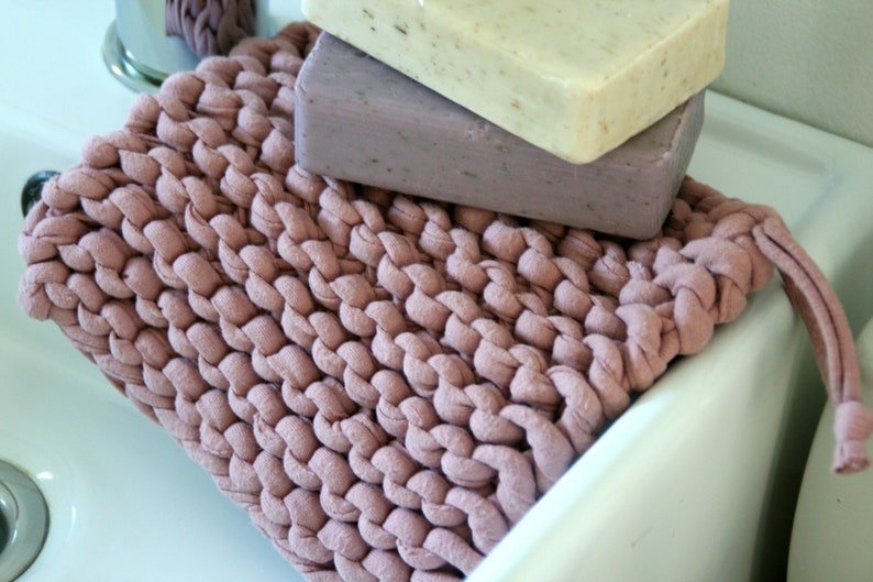 May include: A pink knitted soap dish with three bars of soap on top. The soap dish has a long, thin strap that can be used to hang it up.