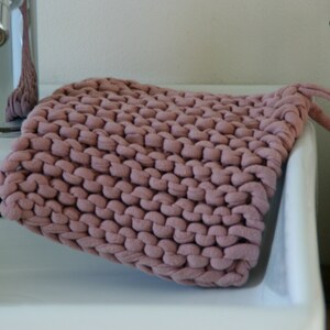 May include: A close-up of a pink knitted dishcloth with a loop handle. The dishcloth is sitting on a white porcelain sink.