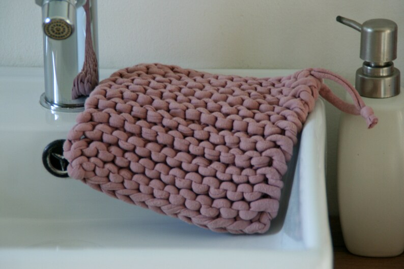 May include: A close-up of a pink knitted dishcloth with a loop handle. The dishcloth is sitting on a white porcelain sink.