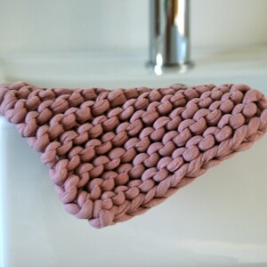 May include: A close-up of a pink knitted washcloth, made from thick yarn, resting on a white porcelain sink.