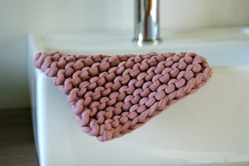 May include: A close-up of a pink knitted washcloth, made from thick yarn, resting on a white porcelain sink.