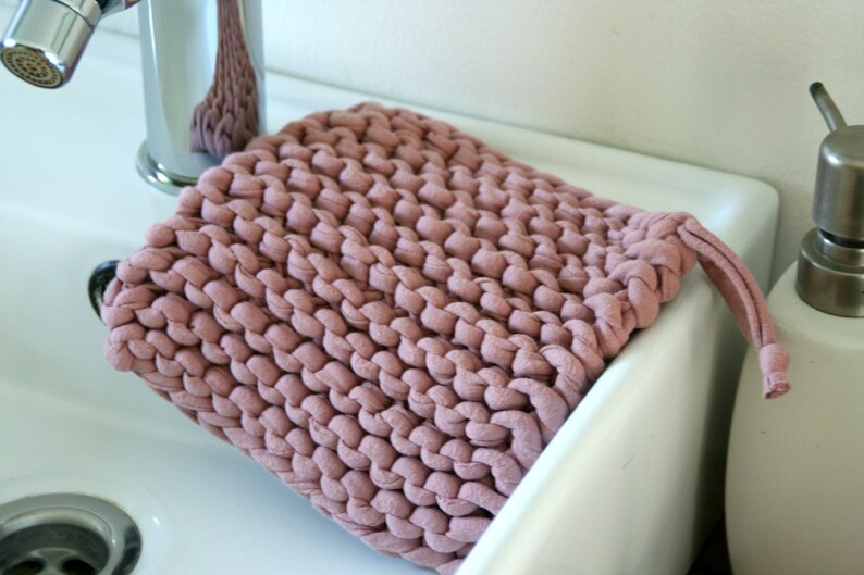 May include: A soft, pink knitted washcloth with a knotted handle. The washcloth is sitting on a white porcelain sink.