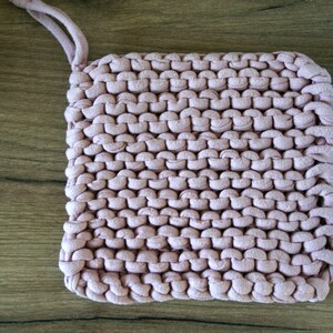 May include: A pink knitted pot holder with a loop handle. The pot holder is made of thick yarn and has a textured surface.