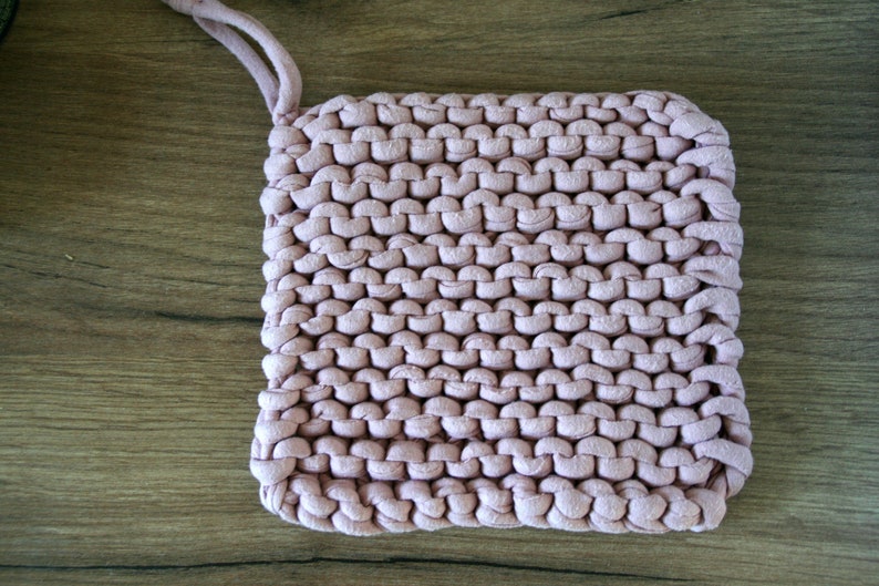 May include: A pink knitted pot holder with a loop handle. The pot holder is made of thick yarn and has a textured surface.