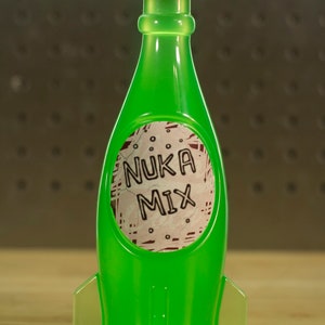May include: A green glass bottle shaped like a rocket with a silver cap. The bottle has a white label with the text "Nuka Mix" in black lettering.