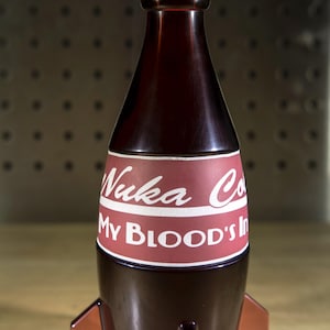 May include: A dark red glass bottle shaped like a rocket with a white label that reads "Nuka Cola" and "My Blood's In It".
