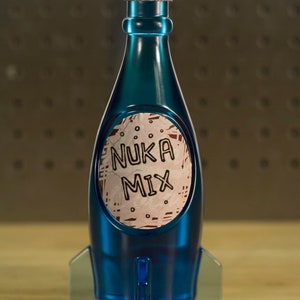 Könnte beinhalten: Eine blaue Glasflasche in Form einer Rakete mit einem silbernen Verschluss. Die Flasche hat ein Etikett mit der Aufschrift "Nuka Mix" in weißen Buchstaben auf rosa-weißem Hintergrund.