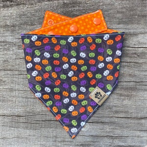 Puede incluir: Bandana reversible para perros con temática de Halloween. Un lado presenta un fondo oscuro con caras de calabaza coloridas. El reverso es naranja con lunares blancos. La bandana tiene un cierre a presión y una pequeña etiqueta con un logotipo de huella de pata.