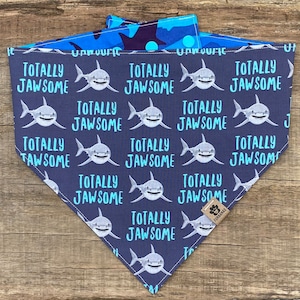 May include: A dark blue bandana with a white shark and the text "Totally Jawsome" repeated in a pattern. The bandana has a blue and white camouflage pattern on the back.