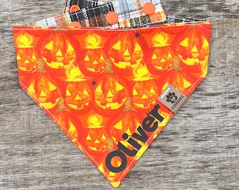 Puede incluir: Un pañuelo reversible para perros con temática de Halloween. Un lado presenta calabazas naranjas con llamas, y el otro lado tiene un patrón de cuadros. El pañuelo está personalizado con el nombre "Oliver" e incluye una etiqueta que dice "Furbanas".
