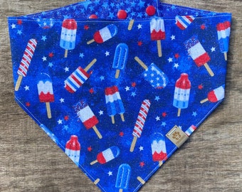 May include: Blue bandana with a red, white, and blue popsicle pattern on a blue background with white stars. The bandana has a brown leather tag with the text "Pawsitive Vibes".