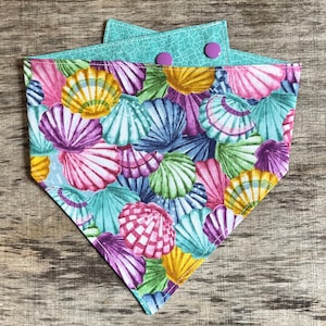 May include: A colorful dog bandana featuring a seashell pattern in shades of pink, purple, yellow, and blue. The bandana has a turquoise backing with two purple buttons for closure. The fabric is a vibrant, eye-catching design.
