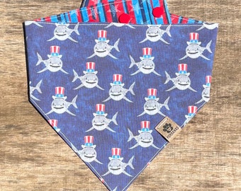 May include: A blue and red dog bandana with a pattern of sharks wearing red, white, and blue patriotic hats. The bandana has a red, white, and blue striped reversible side. The bandana has a small brown tag with the word "FURRIENAS" on it.