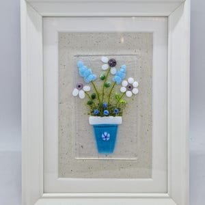 May include: A framed glass art piece depicting a blue flower pot with white and blue flowers. The flowers are made of glass and are arranged in a cluster. The background is a light brown colour.
