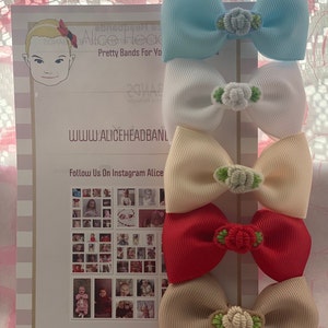 May include: Set of five hair bows in different colours with flower embellishments. The bows are arranged in a stack, with the blue bow on top and the beige bow at the bottom. The bows are made of satin ribbon and have a wired edge. The flowers are made of fabric and have a green stem.