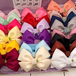 May include: A collection of twenty-four satin hair bows arranged in a box, each featuring a small crocheted rose at the centre. The bows come in a variety of colours including red, pink, yellow, purple, blue, white, black, and green.