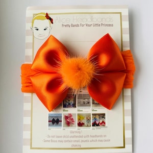 May include: A bright orange satin bow headband featuring a central fuzzy pom-pom accent. The headband is attached to a display card labelled Alice Headbands, Pretty Bands For Your Little Princess, which includes a warning about small parts.