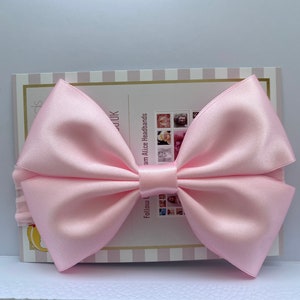 May include: A large pink satin bow headband. The headband is on a white and pink striped background with text that reads "Follow us on Instagram Alice Headbands".