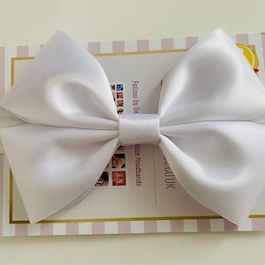 May include: A large white satin bow with a center loop. The bow is on a white and pink striped background with the text "Follow Us On" and "Alice Headbands" in black.