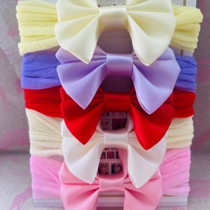 May include: Set of five satin hair bows in various colours, including yellow, lavender, red, white, and pink. The bows are arranged in a stack, with the yellow bow on top and the pink bow on the bottom. Each bow is attached to a stretchy headband.
